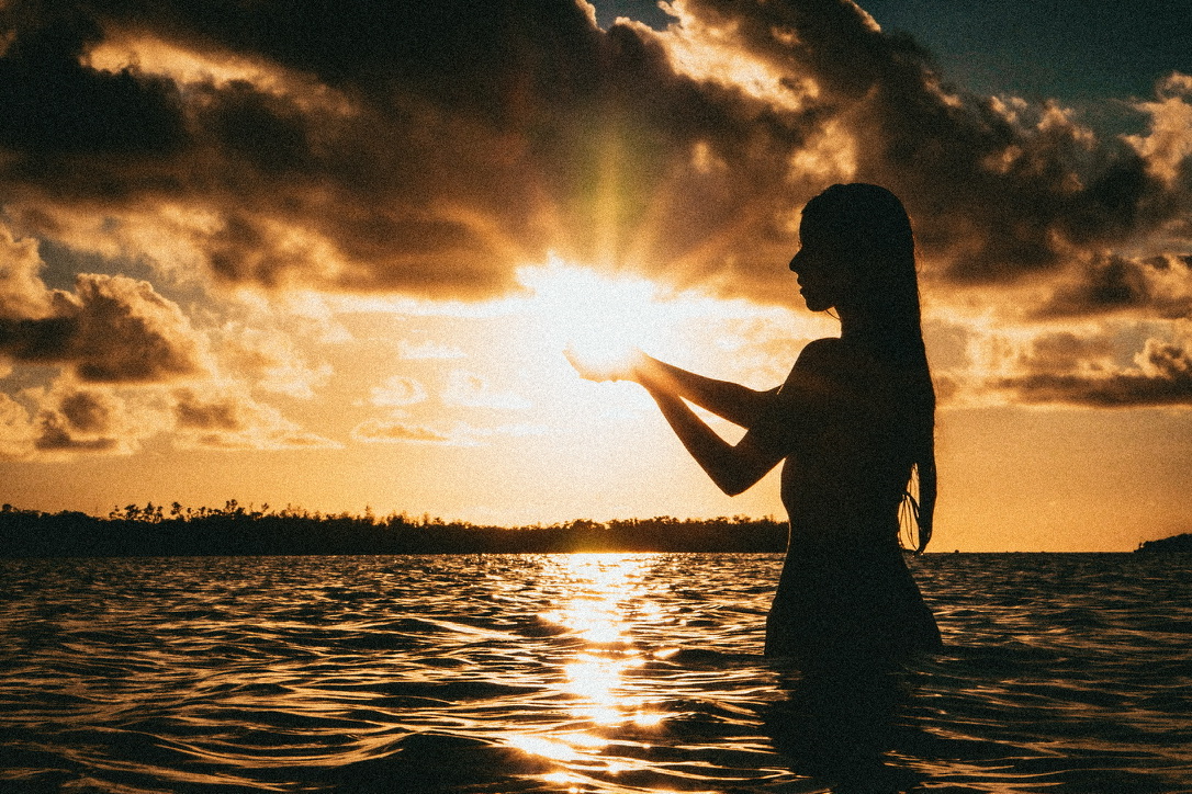 A silhouetted figure holding a pool of sunlight above dark water at sunset